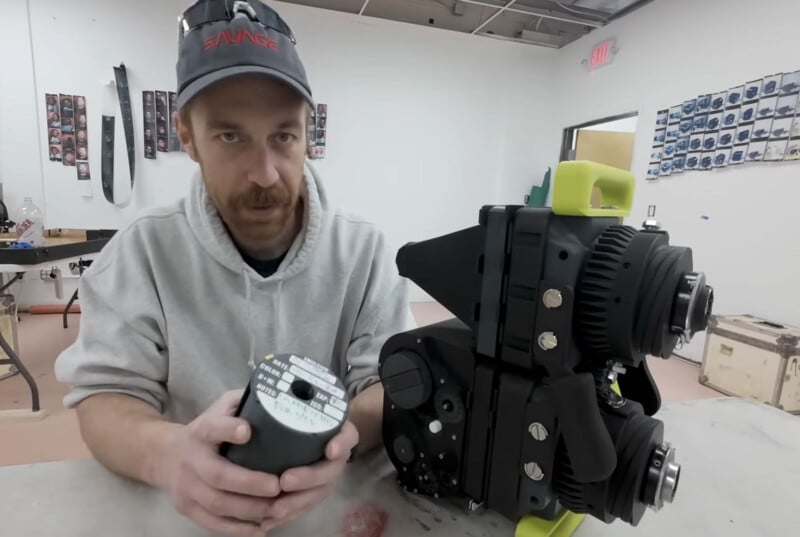 Man in a cap sits at a table holding a part next to a large mechanical device. Walls have numerous small photos. The setting is a workshop with tools, and there's a door in the background.