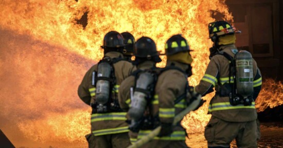 Four firefighters in full gear, including helmets and oxygen tanks, stand closely facing a large, intense blaze. A stream of water can be seen hitting the fire, suggesting they are actively trying to extinguish it.