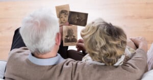 An elderly couple viewed from behind, sitting on a couch and holding old black-and-white photographs. The images in their hands show an infant and a child. The background is a warm, wooden floor.