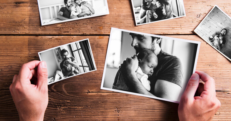 A person holds a black and white photo of a man cradling a baby. Other similar photos are scattered on a wooden surface, depicting intimate moments between a man and a baby.