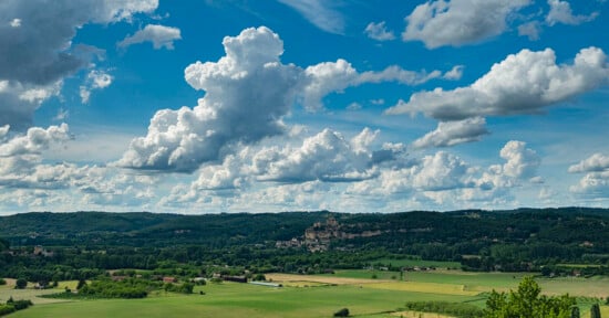 Scenic landscape with a vast green field in the foreground, rolling hills in the background, and a dramatic sky filled with large, fluffy clouds. A small village with clustered buildings is nestled among the hills.