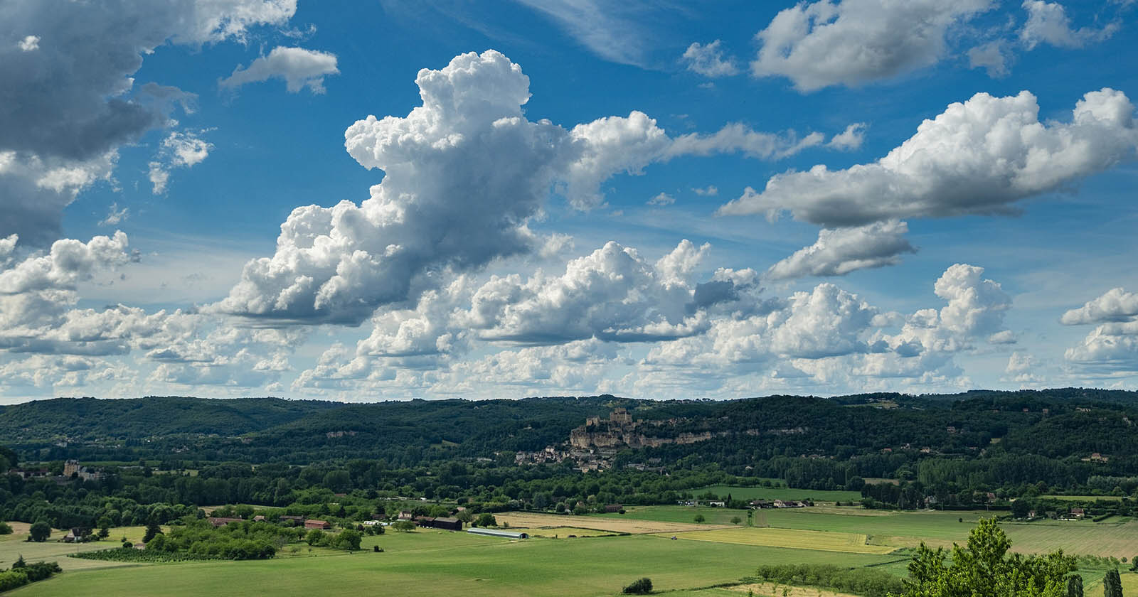 Scenic landscape with a vast green field in the foreground, rolling hills in the background, and a dramatic sky filled with large, fluffy clouds. A small village with clustered buildings is nestled among the hills.