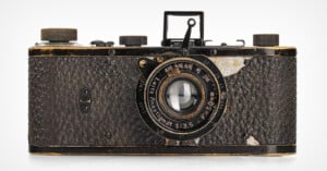 Vintage Leica camera with a worn, textured body and brass accents. The lens reads "Leitz Anastigmat f=5cm 1:3.5" and shows signs of age. The camera has a classic, rectangular design with various dials and a viewfinder on top.