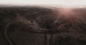 A dramatic aerial view of a vast, deep open-pit mine under a hazy sky. The circular formation is surrounded by winding roads and earth-toned terrain, with distant mountains and a misty horizon in the background.