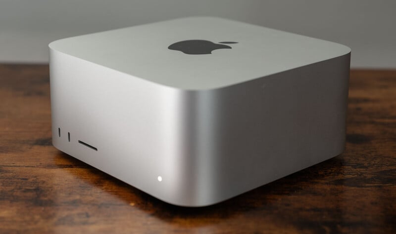 Silver square computer with rounded edges placed on a wooden surface. The device features a black apple logo on top and ports on the front side.