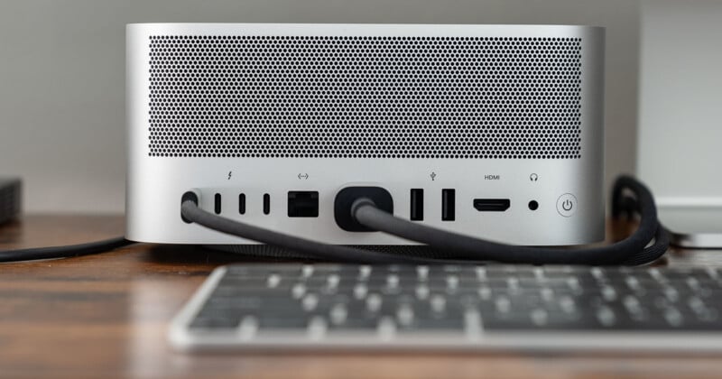 A silver desktop computer with multiple ports, including USB, HDMI, and Ethernet, is situated on a wooden desk. A black cable is connected to one of the ports. A keyboard is partially visible in the foreground.