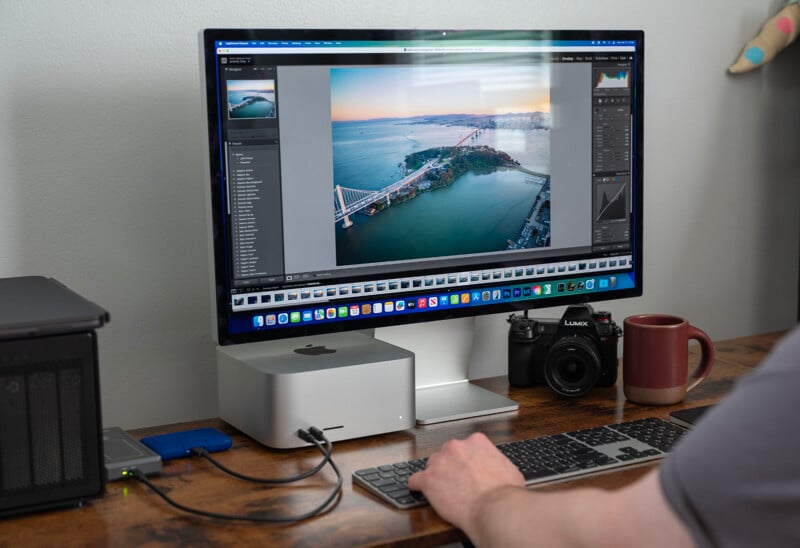 A person working at a desk with a computer displaying a photo editing program. The screen shows an aerial image of a bridge and island. There's a camera, external drive, and mug on the desk.