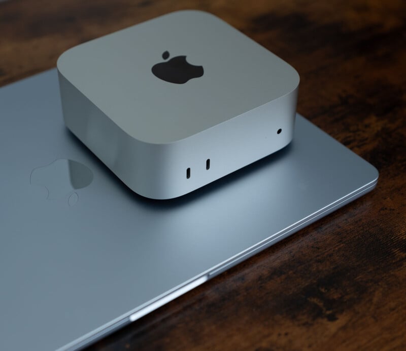 A silver Mac Mini placed on top of a closed silver MacBook, both featuring the Apple logo. The devices are set on a wooden surface.