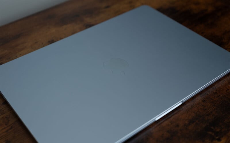 Closed silver laptop on a wooden table, viewed from above. The laptop has a logo in the center of the lid.