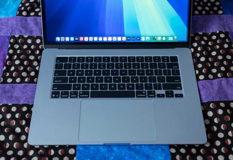 A laptop with a visible keyboard and a screen displaying a blue-themed desktop. The laptop is on a quilt featuring purple, blue, and black patches with a polka dot pattern.