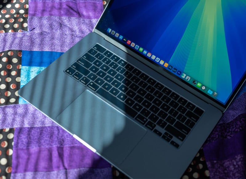 A laptop with a dark keyboard and a colorful screen displaying various icons, resting on a purple and black quilt with circular and geometric patterns. Sunlight creates diagonal shadows across the laptop.