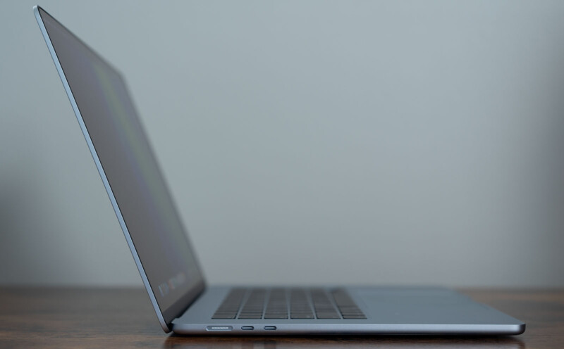 Side view of an open laptop on a wooden surface, displaying a thin profile and minimalist design. The screen is tilted slightly back, and ports are visible on the side. The background is a plain, muted color.