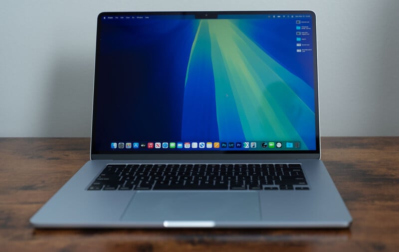 A laptop with a dark keyboard is open on a wooden surface. The screen displays a blue and green abstract wallpaper with app icons lined up at the bottom. The desktop shows some icons on the right side.