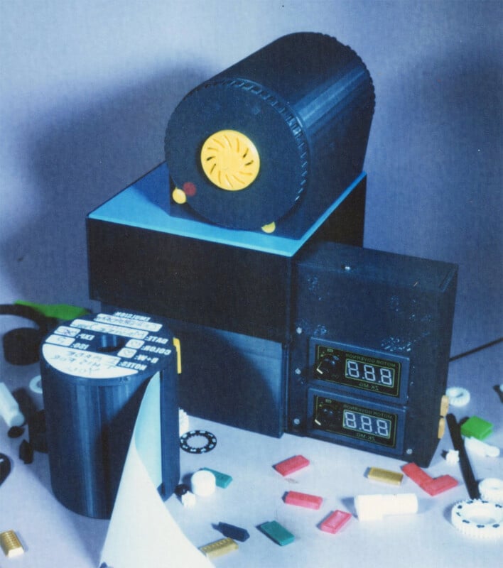 A vintage electronic device with a cylindrical component on top and digital displays on the front. Various small colorful objects and gears are scattered around. The setup is placed on a neutral surface.