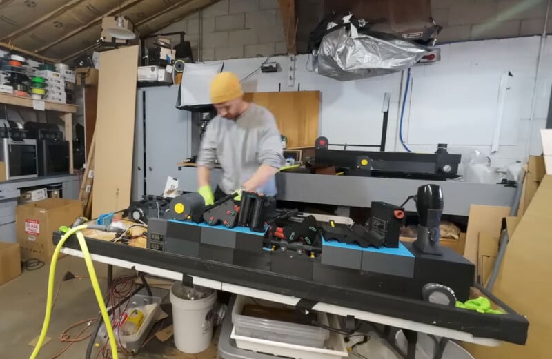 A person wearing a yellow beanie and gray sweater works at a cluttered workbench in a garage or workshop. The table is filled with various tools and equipment. Shelving and storage boxes are visible in the background.