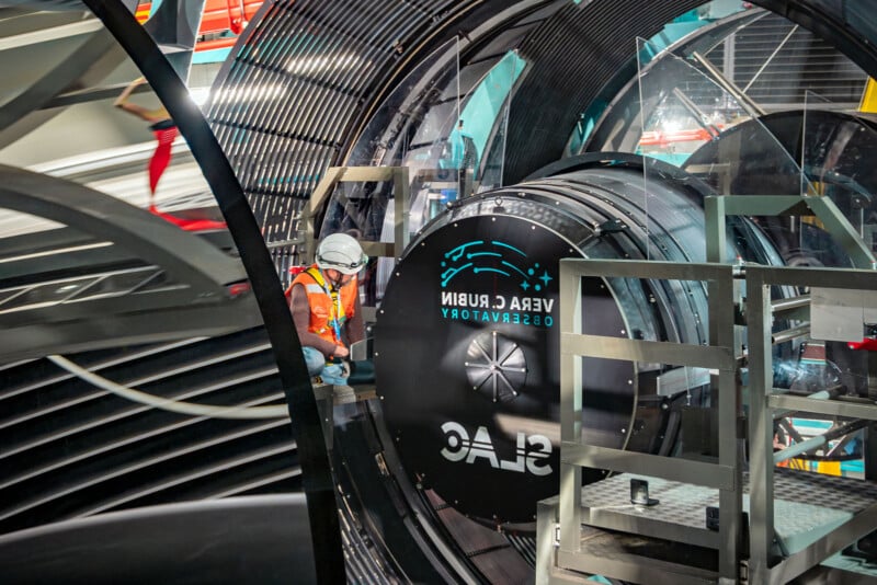 A person wearing a hard hat and safety vest inspects equipment inside a large cylindrical structure. The equipment displays "Vera Rubin Observatory" and "SLAC" logos. The setting is industrial, with metallic and mechanical components.