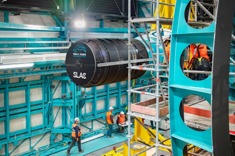 A large black cylindrical component labeled "SLAC" and "Vera C. Rubin Observatory" is being maneuvered by a crane inside an industrial facility. Workers in orange vests and hard hats are guiding the process amidst scaffolding and metal structures.