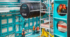 Workers in hard hats and safety gear assemble a large black scientific instrument labeled "Vera C. Rubin Observatory" and "SLAC" inside a blue and white industrial facility. Scaffolding and equipment surround the area.