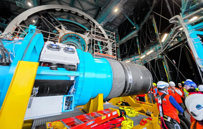 A group of construction workers wearing helmets and safety vests stand around a large piece of industrial machinery inside a facility. The machinery is blue and yellow, with a cylindrical metallic component. Scaffolding and structures are visible in the background.