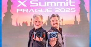 Two men standing in front of a backdrop reading "X Summit Prague 2025" hold cameras towards the viewer. The backdrop shows a silhouette of a cityscape at sunset. Both men are smiling and dressed casually.