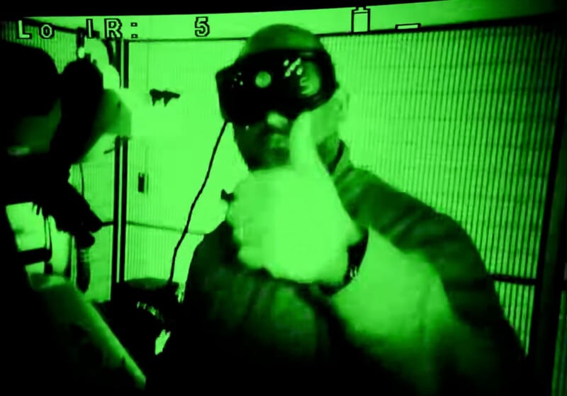 A person wearing night vision goggles gives a thumbs-up gesture. The image is seen through a green-tinted night vision filter, creating a glowing effect. Equipment or machinery is partially visible beside the person.