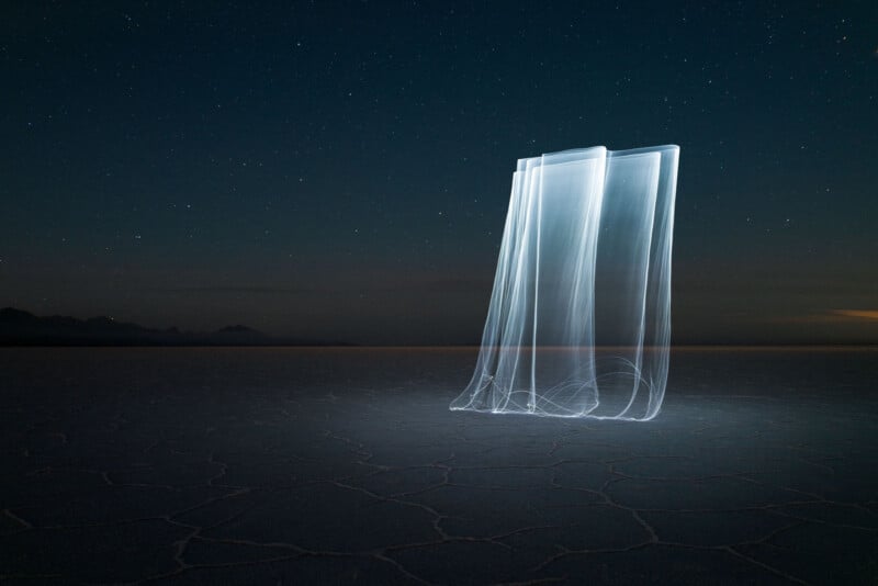 A long-exposure photograph of a starry night sky over a vast, flat landscape with hexagonal patterns on the ground. In the foreground, ethereal light trails create vertical, curtain-like shapes glowing white against the dark backdrop.