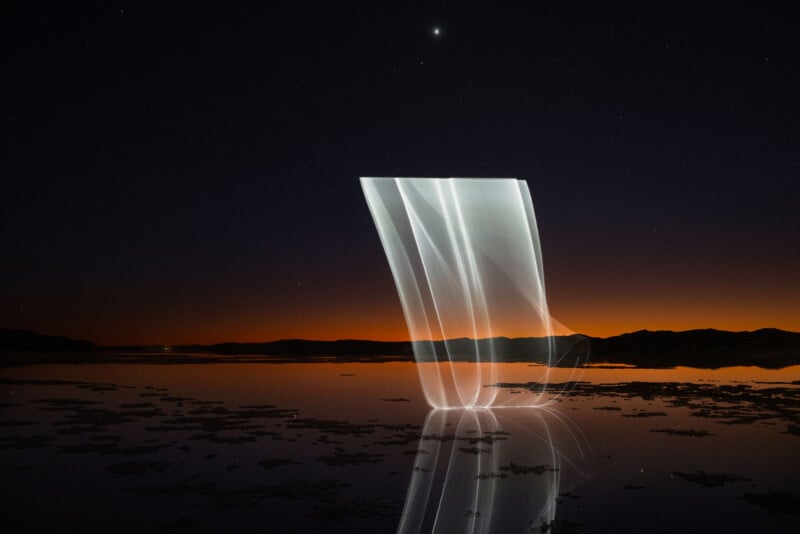 A serene night scene by a lake with a starry sky. Soft, flowing light trails form graceful curves reflecting on the water's surface, contrasting against a deep orange horizon with silhouetted hills.