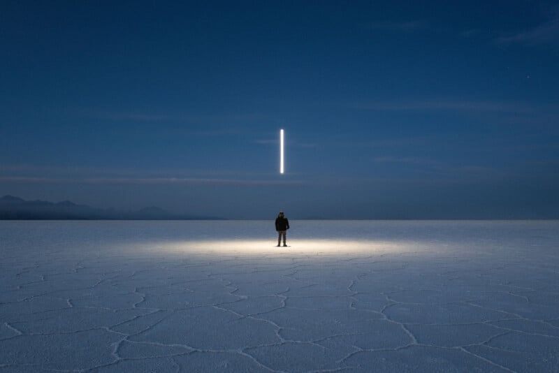 A person stands on a vast, cracked white surface under a deep blue sky. A bright vertical light hovers above them, casting a glow on the ground. Mountains are faintly visible in the background.