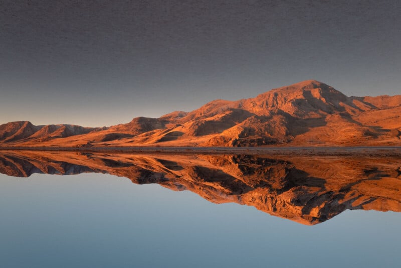 Panoramic view of a mountain range bathed in warm, golden sunlight at sunset. The mountains are perfectly reflected in the still, mirror-like water below, creating a symmetrical and serene landscape scene.