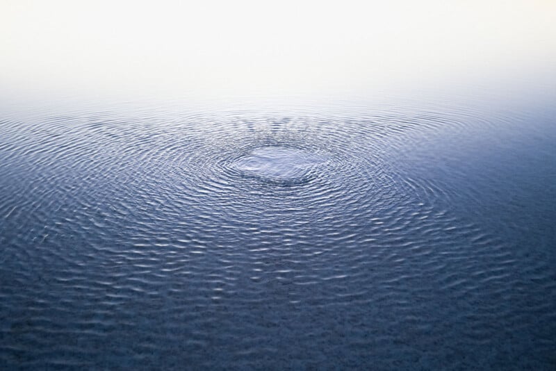 Superfície de água calma com ondulações concêntricas em expansão de um ponto central, criando um efeito visual sereno e pacífico. A água reflete um céu claro, aumentando a tranquilidade da cena.