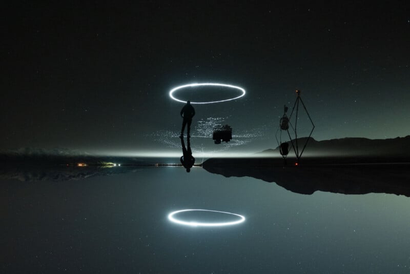 Two silhouettes stand on a reflective surface under a starry night sky. One person holds a hoop of light, creating a halo effect mirrored below. Mountains and distant lights are visible on the horizon, enhancing the surreal ambiance.