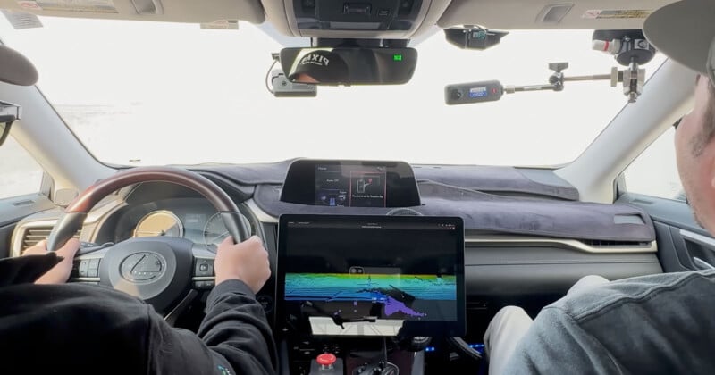 Interior of a car from the back seat view, with two people in the front. The driver is holding the steering wheel. A large monitor displays colorful graphs, possibly a mapping system. Various devices are mounted on the dashboard and windshield.