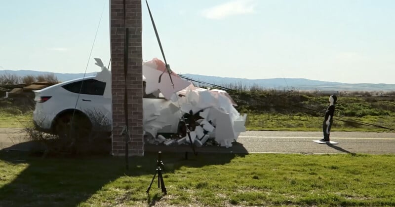 A white car crashes into a brick wall covered with foam blobs, creating an explosion of foam. A crash test dummy stands on the road nearby. A grassy field and mountains are in the background under a clear sky. Two tripods are on the grass.