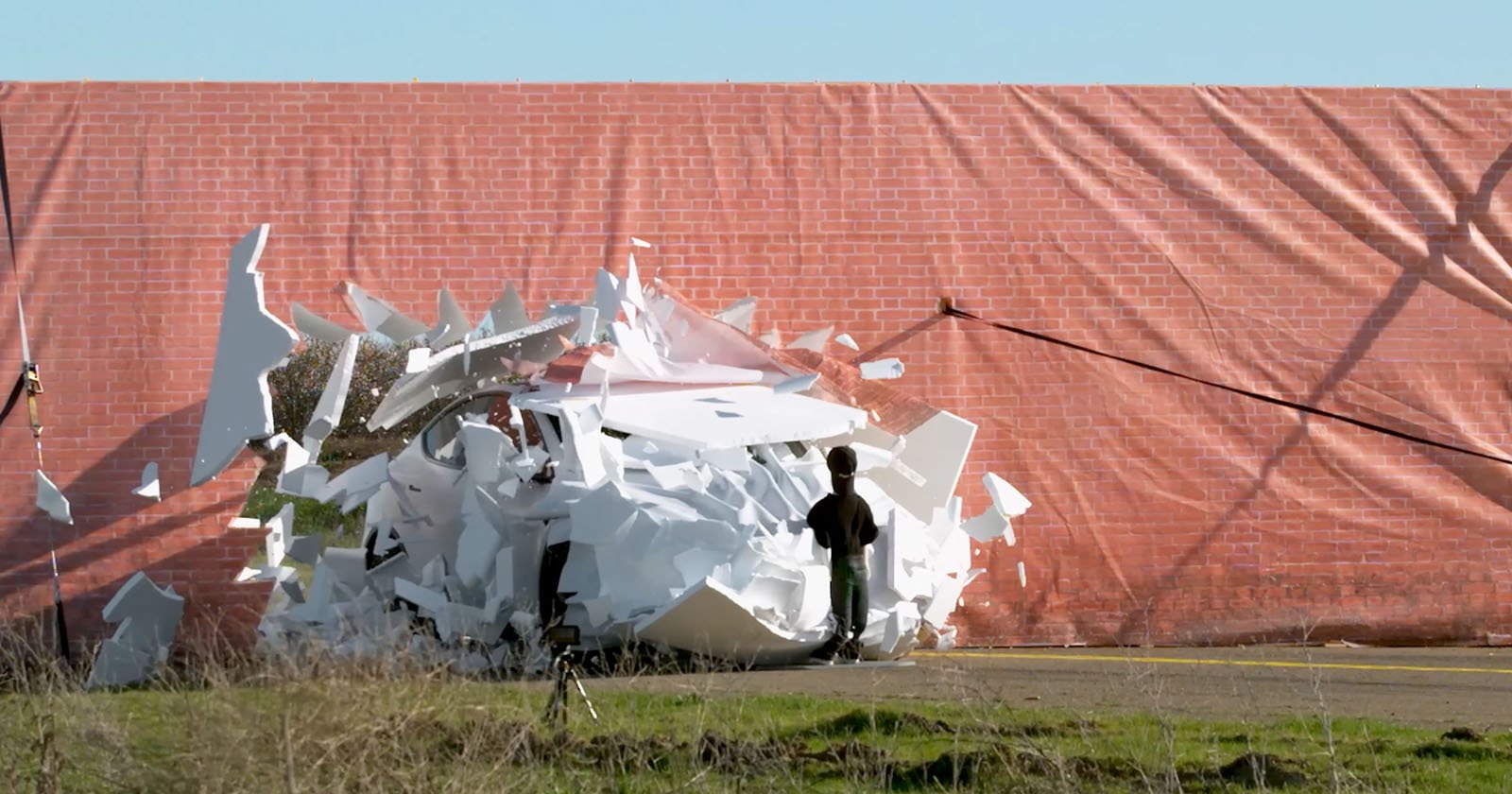 A white car is dramatically crashing through a fake brick wall made of lightweight materials, creating a dynamic spray of debris. A person stands nearby, watching the action. The scene is set outdoors with grass and a clear sky.