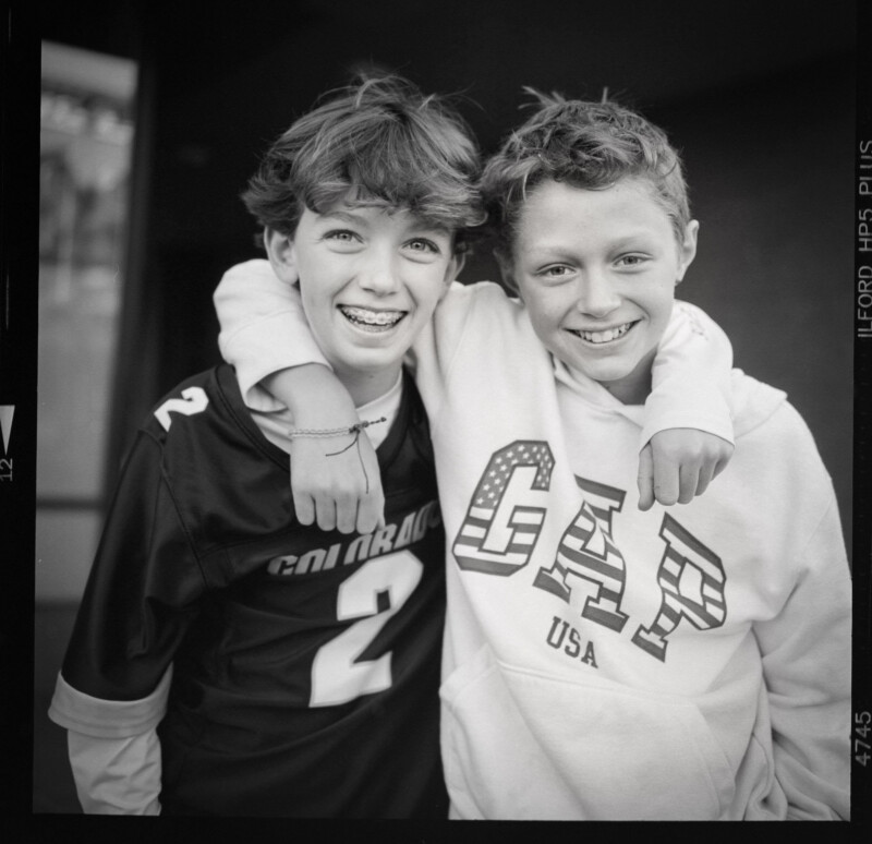 Two smiling boys stand close, one wearing a sports jersey with the number 2 and the other in a GAP USA hoodie. The boy in the jersey has braces, and they both have their arms around each other, conveying friendship. The image is in black and white.