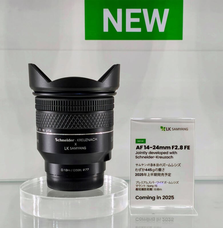 A camera lens labeled "Schneider - Kreuznach x LK Samyang AF 14-24mm F2.8 FE" is displayed on a clear stand. Next to it is a sign with the same details and the note, "Coming in 2025," beneath a green "NEW" sign.