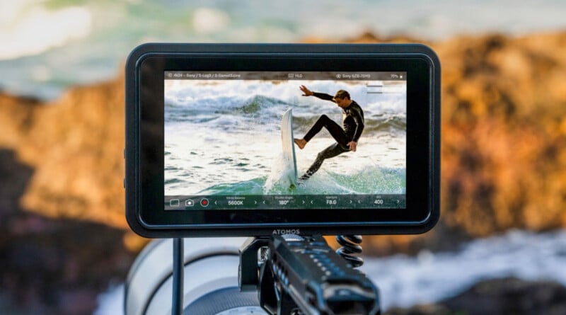 A camera monitor displays a surfer performing a jump on a wave. The image captures the action in sharp detail. The background shows a blurred view of the ocean and rocks.