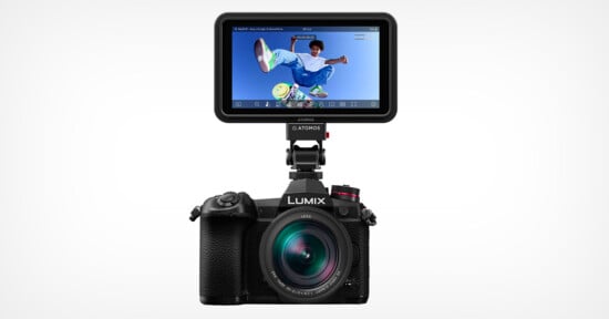 A black Lumix camera with a lens facing forward is topped with an Atomos external monitor displaying an image of a skateboarder mid-air against a clear blue sky. The setup is on a plain white background.