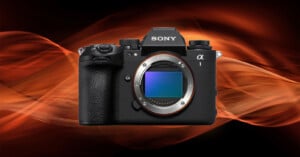 A Sony Alpha camera body with the lens removed, revealing the sensor, is displayed against a dynamic background of flowing orange streaks on a dark backdrop.