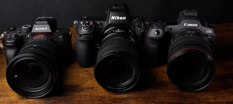 Three cameras are lined up on a wooden surface against a dark background. From left to right, they are a Sony, Nikon, and Canon. Each camera has a lens attached.