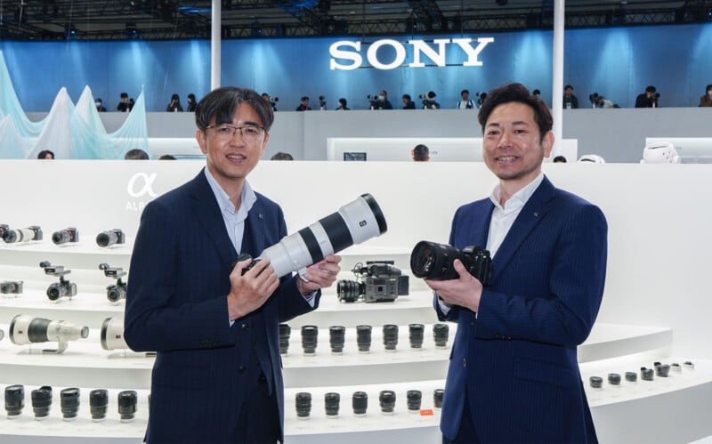 Two men in suits stand in front of a Sony display. One holds a large telephoto lens, and the other holds a camera and lens. Behind them are rows of camera lenses and equipment under the Sony logo.