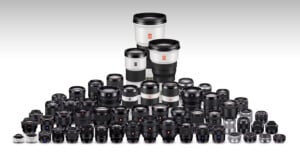 A large collection of camera lenses is arranged in rows. They vary in size and type, including wide-angle, telephoto, and prime lenses, with a few larger white lenses in the back. The background is a light gray gradient.