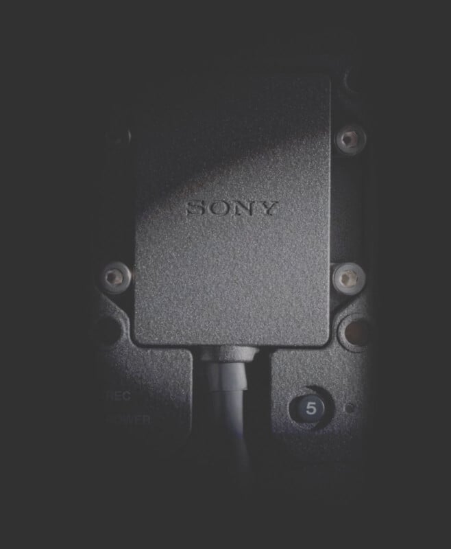 Close-up of a textured, rectangular Sony device with cables attached at the bottom. The device is surrounded by a dark background. There's a visible label with the number 5 near the bottom right corner.