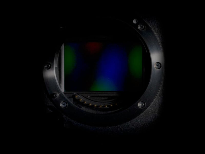 Close-up view of a camera sensor with a multicolored, abstract glow on its surface. The image is framed by a dark, circular lens mount, highlighting the electronic connections. The background is dark, emphasizing the sensor's vibrant colors.