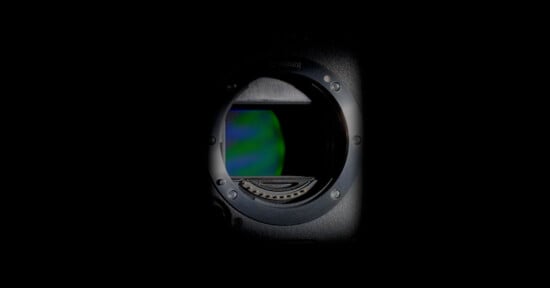 Close-up of a camera lens opening with visible sensor inside, surrounded by a dark background. Light reflects off the inner metal ring, highlighting the sensor's reflective surface.