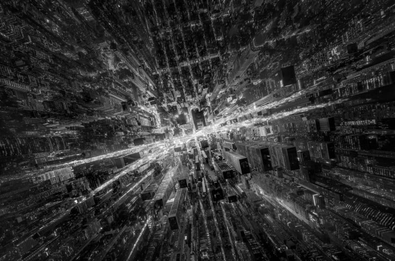 Aerial black and white view of a densely packed cityscape at night. Skyscrapers and streets are illuminated, creating a dynamic, radiating pattern of lights that appear to converge towards the center, giving a sense of depth and movement.