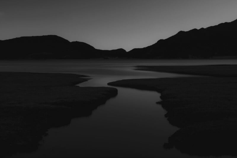 A serene landscape with a winding river flowing through a dark, open field at dusk. Silhouetted hills and mountains frame the horizon under a dimly lit sky, creating a peaceful, reflective atmosphere.