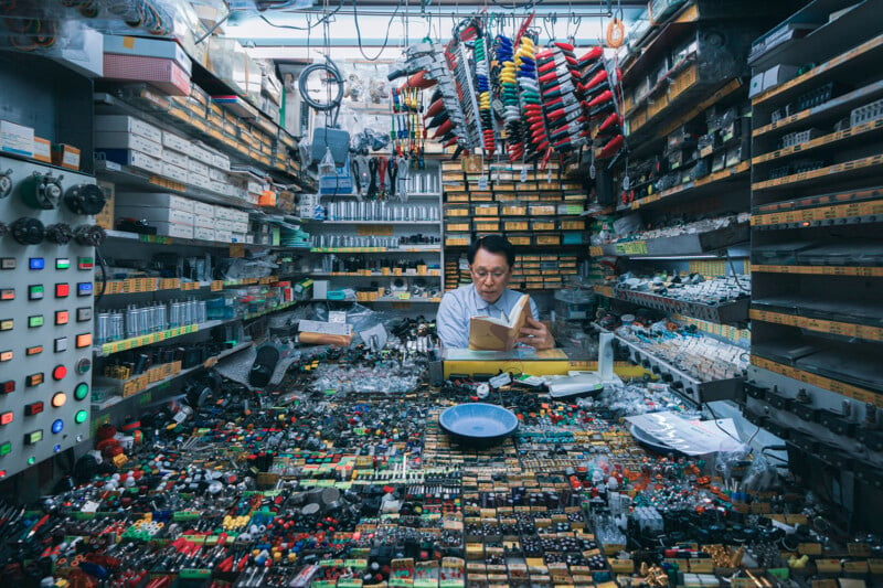 Man sits reading a book in a cluttered electronics store filled with shelves of parts, colorful wires, and gadgets. Numerous drawers and switches cover the walls. The space is packed with electronic components and tools.