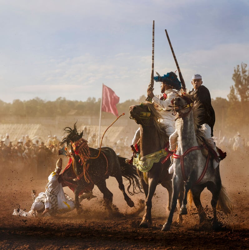 Horsemen in traditional attire ride through a dusty field during a cultural event. One horse appears to be bucking, and a rider is falling to the ground. A pink flag is visible in the background, and spectators are watching.