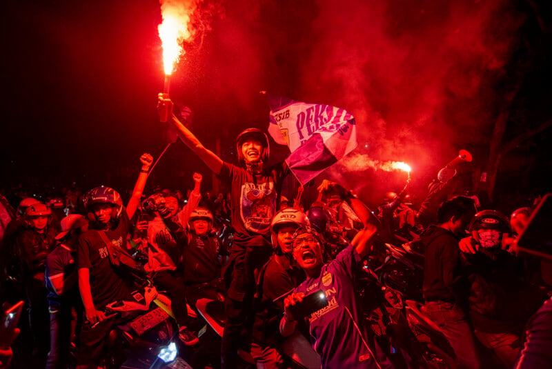 A group of people, some on motorcycles, celebrate energetically at night, holding flares and flags. The scene is lit with red light and filled with smoke, creating a vibrant and dynamic atmosphere.